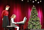 Dziewczynka w kręconych włosach na pianinie elektronicznym, za nią stoi kobieta w czerwonym swetrze, z drugiej strony choinka