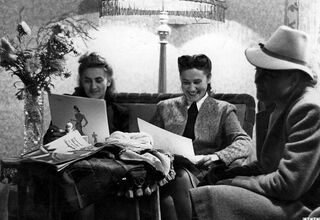 Zdjecie czarno-białe. Trzy eleganckie kobiety, jedna w kapeluszu, siedzą przy stole i oglądają katalogi mody