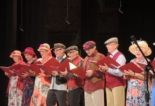 Kobiety i mężczyźni w kolorowych strojach śpiewają na scenie.