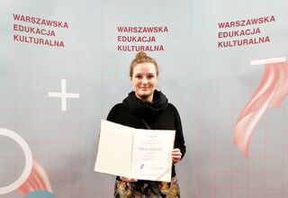 Katarzyna Anuszewska prezentuje dyplom na tle ścianki Warszawskiej Nagrody Edukacji Kulturalnej