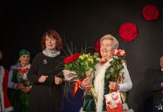 Dwie kobiety, jedna w czarnej sukience, druga trzyma bukiet kwiatów i torebki prezentowe  i uśmiecha się