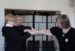 Trzy kobiety ubrane na czarno i lekko uśmiechnięte ciągną się za ręce