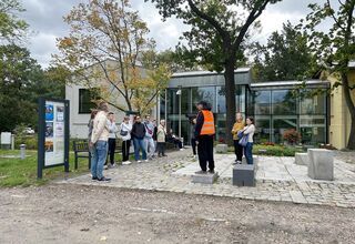 Grupa osób z przewodnikiem w pomarańczowej kamizelce, w tle szklany budynek