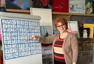 Zdjęcie: Uśmiechnięta kobieta-seniorka przy flipcharcie z grą sudoku, w tle kolorowe obrazy