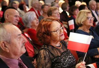 Zdjęcie: grupa seniorów w odświętnych strojach, na pierwszym planie seniorka z biało-czerwoną flagą