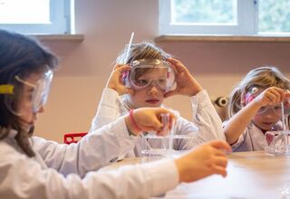 Zdjęcie: troje dzieci siedzących przy stole w białych fartuchach i okularach ochronnych