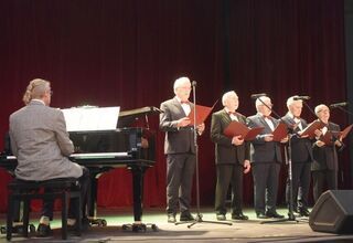 Zdjęcie mężczyzn w ciemnych garniturach śpiewających na scenie. Mężczyzna w szarej marynarce grający na pianinie