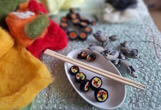 Zdjęcie: filcowe sushi w białym naczyniu, nad nimi drewniane pałeczki, z prawej strony kilka filcowych myszek, po lewej stronie kolorowy filc