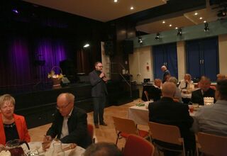 Zdjęcie mężczyzny trzymającego mikrofon, obok seniorzy siedzący przy stołach wielkanocnych