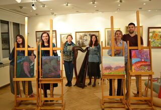 Zdjęcie: grupa czterech młodych osób stojących przy sztalugach z obrazami, z tyłu dwie kobiety przy harfie, w tle obrazy na ścianie