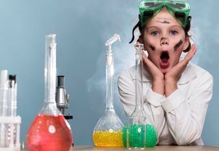 Zdjęcie: Dziewczynka w białym fartuchu i zielonych okularach ochronnych na głowie ze zdziwioną miną podpierająca się rękami na stole, na pierwszym planie naczynia z kolorowymi substancjami