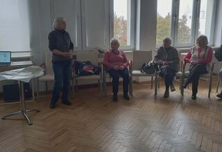 Zdjęcie stojącej starszej kobiety w otoczeniu seniorek siedzących na krzesłach