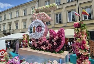 Zdjęcie: instalacja artystyczna z kwiatów, w jasnych kolorach, symbolizująca warszawską syrenkę, w tle kamienice