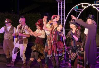 Zdjęcie: grupa osób w steampunkowych kostiumach na scenie, fioletowe oświetlenie, obok metalowa konstrukcja z zegarem,