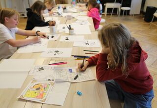 Zdjęcie: dzieci stojące przy podłużnym stole, na nim kartki, kredki i cienkopisy