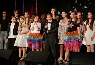 Zdjęcie: dzieci i młodzież na scenie, w kolorowych ubraniach, trzy dziewczynki z mikrofonami w ręku