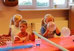 Troje dzieci siedzi przy stole. Dziewczynka dmucha balony.