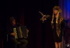 Kobieta w szalu, z futrzaną czapką i ptakiem na ramieniu występuje na scenie. Obok na akordeonie gra mężczyzna.