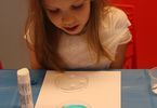 Dziewczynka siedzi przy stole i patrzy na kartkę, na której położony jest pojemnik z niebieskim płynem i gwiazdkami
