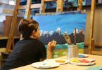 Dziecko maluje obraz. Obok niego znajduje się stół z przyborami do malowania.