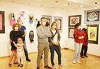 Grupa ludzi stoi na tle wystawy obrazów i materiałowych lalek.