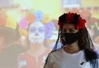 Kobieta z welonem i sztucznymi kwiatami na głowie stoi na tle zdjęcia kobiety w makijażu z okazji Día de los Muertos.