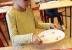 Dziewczynka niosąca papierowy talerzyk z brokatem. Przed nią stoi stół z ozdobami.