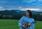 Uśmiechnięta dziewczyna grająca na ukulele. Stoi na łące, na tle gór.