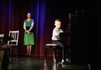 Chłopiec gra na pianinie, w tle stoi instruktorka i przygląda się chłopcu.