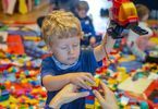 Chłopiec trzymający wysoko uniesioną rękę, a w niej helikopter, częściowo wykonany z klocków Lego, który drugą ręką przytrzymuję budowlę w kształcie wieży z klocków Lego