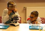 Dwóch chłopców jeden stoi i patrzy przez mikroskop drugi siedzi uśmiechnięty