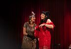 Dwie kobiety ubrane  eleganckie wieczorowe sukienki śmieją się, w tle ciemnoczerwona kotara