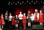Dzieci stoją na scenie w strojach świątecznych, za nimi kobieta mówi do mikrofonu