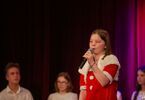 Dziewczynka w świątecznej sukience śpiewa do mikrofonu, w tle młodzież