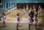 Dziewczynki w strojach baletowych stoją bokiem do instruktorki, w tle lustro