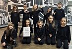 Zdjęcie: zespół dziecięcy w czarnych strojach, część osób podparte na kolanach, za nimi osoby stojące, jedna dziewczynka trzyma dyplom