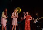 Zdjęcie: trzy młode dziewczyny w sukienkach z mikrofonami w ręku, jedna z nich śpiewająca, w tle zespół muzyczny, lampa w żółtym kolorze i bordowa kotara