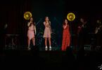 Zdjęcie: trzy śpiewające młode dziewczyny w kolorowych sukienkach z mikrofonami w ręce, w tle zespół muzyczny, dwie lampy w żółtym kolorze i bordowa kotara