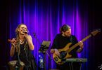 Zdjęcie: wokalistka i gitarzysta na scenie, w tle kotara podświetlona na fioletowo