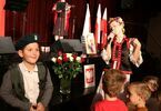 Zdjęcie: dzieci przy scenie strojące obok kobiety z mikrofonem w tradycyjnym stroju ludowym, chłopiec z wojskową czapką i plecakiem, na scenie młoda kobieta przy mikrofonie, z przodu róże w kolorze białym i czerwonym w wazonie oraz godło Polski