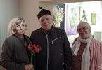 Zdjęcie: trzy stojące osoby, mężczyzna w kurtce i nakryciu głowy w środku, po jego obu stronach dwie seniorki, jedna z nich trzymająca kwiat w dłoni