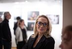 Zdjęcie: uśmiechnięta kobieta w okularach w czarnej marynarce na tle grupy osób i wiszących na ścianie fotografii