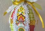 Pisanka w żółto-białej kolorystyce z czerwonymi akcentami ozdobiona kolorowymi skrawkami tkanin, koronką, wstążkami oraz koralikami