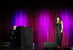 Zdjęcie: kobieta w średnim wieku na scenie z mikrofonem w czarnym kombinezonie, po lewej stronie pianino, w tle kotara w fioletowo-różowym odcieniu
