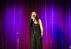 Zdjęcie: kobieta w średnim wieku na scenie z mikrofonem na fioletowym tle