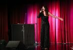 Zdjęcie: kobieta w średnim wieku na scenie z mikrofonem, po lewej stronie pianino, w tle kotara w czerwonym odcieniu