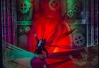 Zdjęcie: postać z rozłożonymi rękoma w steampunkowej scenografii w czerwono-czarnej kolorystyce