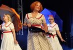 Zdjęcie: panie w dojrzałym wieku na scenie w sukienkach w kropki z kolorowymi parasolkami nad głowami, z przodu kobieta ze złożoną parasolką