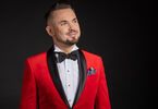 Zdjęcie: lekko uśmiechnięty mężczyzna w eleganckiej czerwonej marynarce smokingowej z czarnymi klapami, białej koszuli i czarnej muszce na ciemnym tle, w kieszonce marynarki kolorowa poszetka