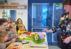 Zdjęcie: dzieci z opiekunkami przy stoliku wykonujące prace plastyczne, po prawej stronie kobieta widziana z profilu pokazująca palcem na papierowe kwiaty